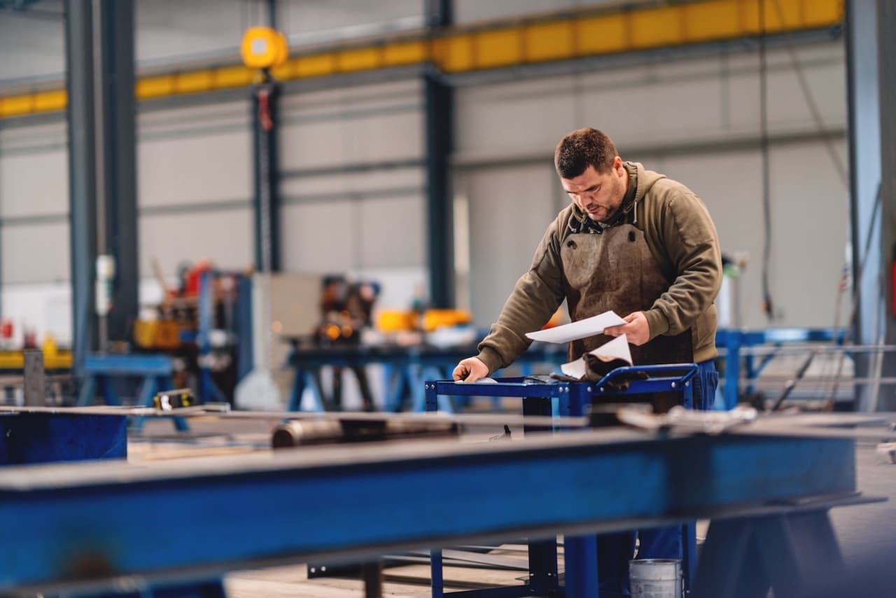 Trabajador operando maquinaria en una fábrica, con un ventilador TBSI-580 en el fondo, asegurando un ambiente laboral cómodo.